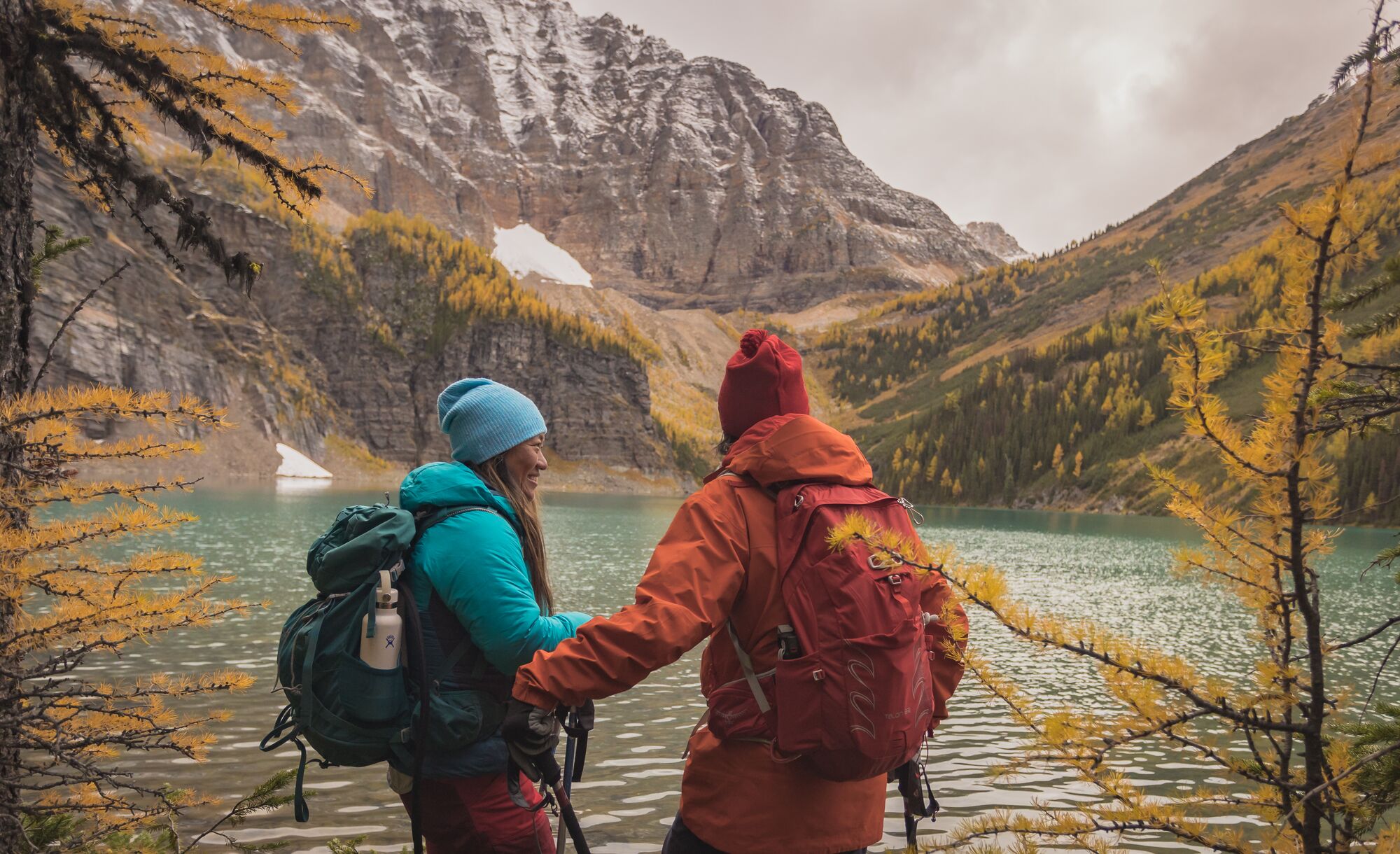 A man and woman hike alongside a lake with yellow larch trees and fresh snow on the mountains surrounding them